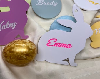 May include: A set of Easter decorations featuring white bunny shapes with names written on them, including Emma, Haley, Brody, and Robert. There is also a gold-colored Easter egg and a light blue egg.