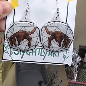 May include: A pair of dangle earrings featuring a clear, geometric, multi-faceted shape with a brown, multi-legged creature illustration inside. The earrings are attached to a white display card with the text HANDMADE and SIGHTLY ART.