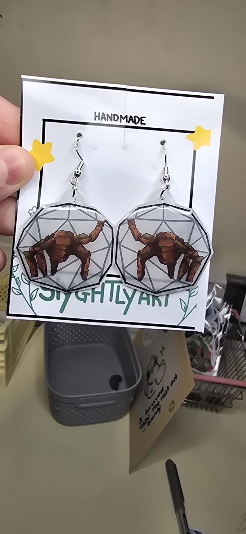 May include: A pair of dangle earrings featuring a clear, geometric, multi-faceted shape with a brown, multi-legged creature illustration inside. The earrings are attached to a white display card with the text HANDMADE and SIGHTLY ART.
