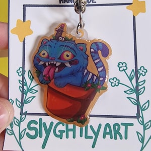 May include: A blue cat keychain with a red tongue sticking out, sitting in a red pot. The keychain has a silver clasp and is attached to a card with the text "Slyghily Art".