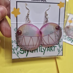 May include: Handmade earrings with a cartoon character design. The earrings are round, with a pink and blue gradient, and feature a smiling face with sharp teeth. The text "Slyghtly Art" is visible. The card reads "HANDMADE".