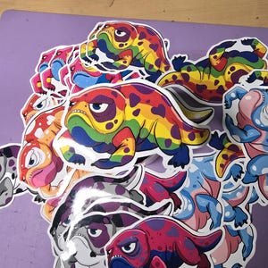 Puede incluir: Una colección de pegatinas coloridas con criaturas de estilo de dibujos animados con manchas de colores arcoíris. Las criaturas tienen una expresión gruñona y están en varias poses.