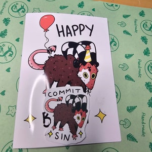 May include: Two stickers featuring a cartoon opossum with a red nose and large eyes. The first sticker shows the opossum wearing a party hat and holding a balloon. The text "HAPPY" is above the opossum. The second sticker shows the opossum wearing a striped hat and the text "COMMIT TO SIN".