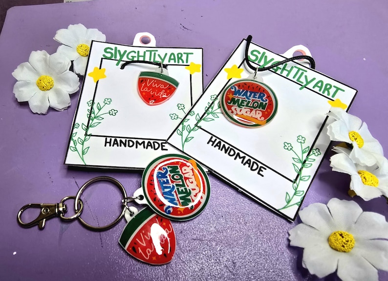 May include: Handmade watermelon-themed jewelry and keychain set. The set includes a pendant with "Viva la Vida" and a watermelon slice design, and a pendant with "Watermelon Sugar". The keychain has a round pendant and a watermelon slice pendant.
