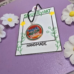 May include: A handmade necklace with a round pendant featuring the words "Watermelon Sugar" in red and blue. The pendant hangs from a black cord and is displayed on a white card with the text "Handmade" and "Slightly Art". The background is purple with white flower accents.
