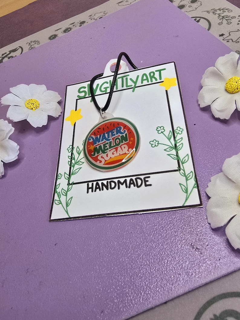 May include: A handmade necklace with a round pendant featuring the words "Watermelon Sugar" in red and blue. The pendant hangs from a black cord and is displayed on a white card with the text "Handmade" and "Slightly Art". The background is purple with white flower accents.