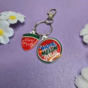 May include: A keychain with two charms. One charm is a red watermelon slice with the words "Viva la vida" in white. The other charm is a round watermelon with the words "Watermelon Sugar". The keychain has a silver clasp and ring.