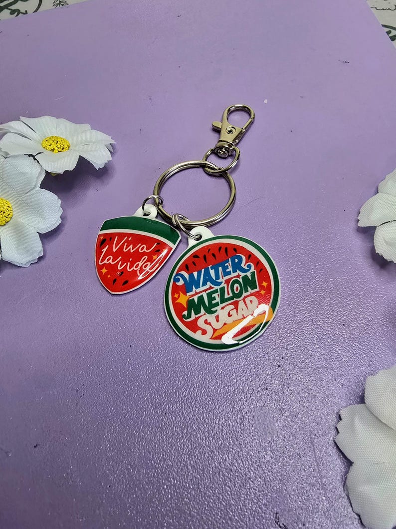 May include: A keychain with two charms. One charm is a red watermelon slice with the words "Viva la vida" in white. The other charm is a round watermelon with the words "Watermelon Sugar". The keychain has a silver clasp and ring.