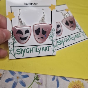 May include: Handmade earrings depicting theatre masks, one smiling and one crying, in pink and red hues. The earrings are presented on a white card with the text "Slyghtly Art" and the word "Handmade".