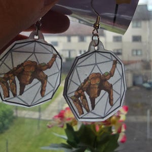 May include: A pair of dangle earrings featuring a flat, translucent, hexagonal charm with a geometric line pattern and a brown, multi-limbed creature illustration. The earrings are attached to a white display card with the partial text ADE.