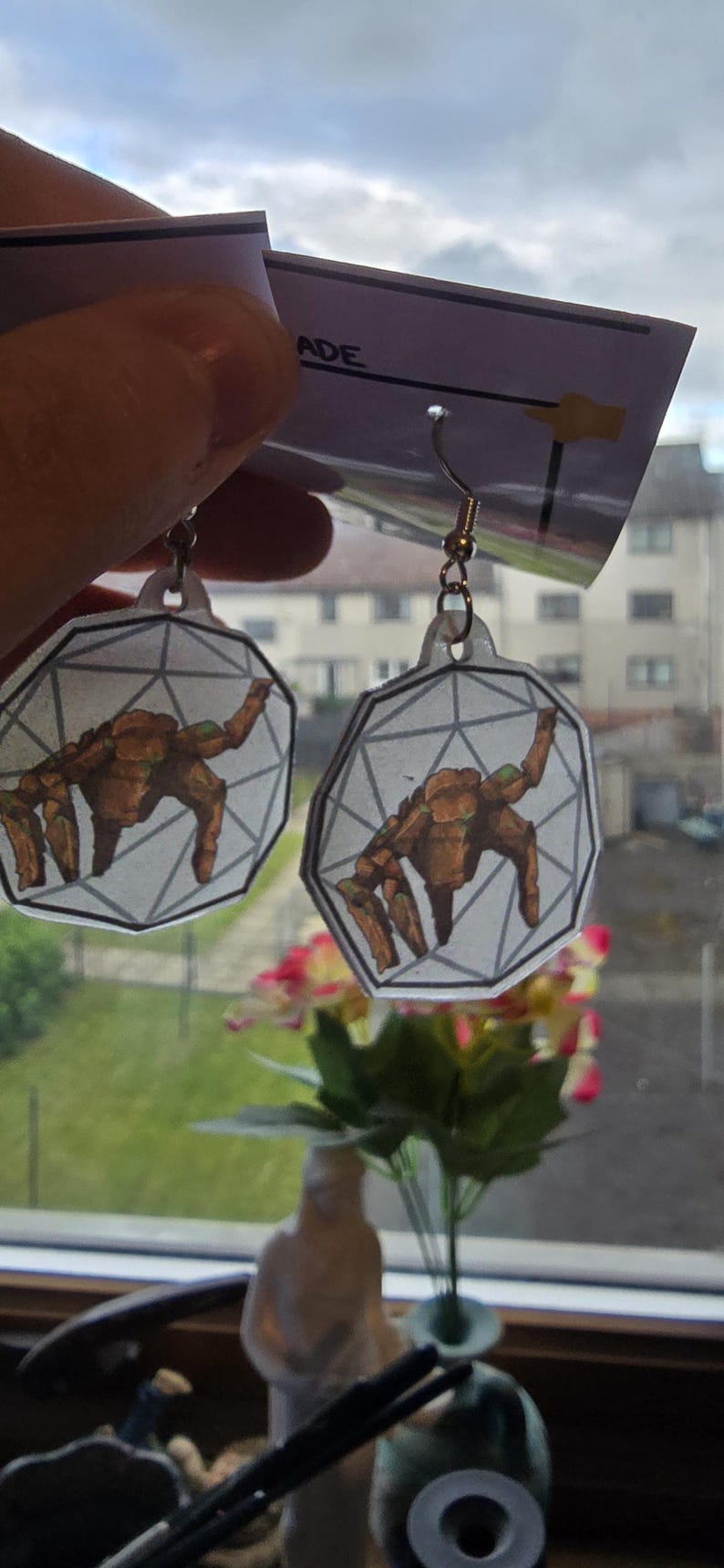 May include: A pair of dangle earrings featuring a flat, translucent, hexagonal charm with a geometric line pattern and a brown, multi-limbed creature illustration. The earrings are attached to a white display card with the partial text ADE.