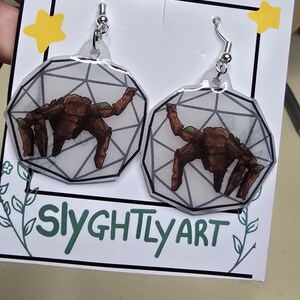 May include: A pair of dangle earrings featuring a brown, multi-legged creature illustration inside a geometric, translucent, white, dodecahedron-shaped frame. The earrings are attached to a white display card labeled HANDMADE and SLYGHTLY ART.