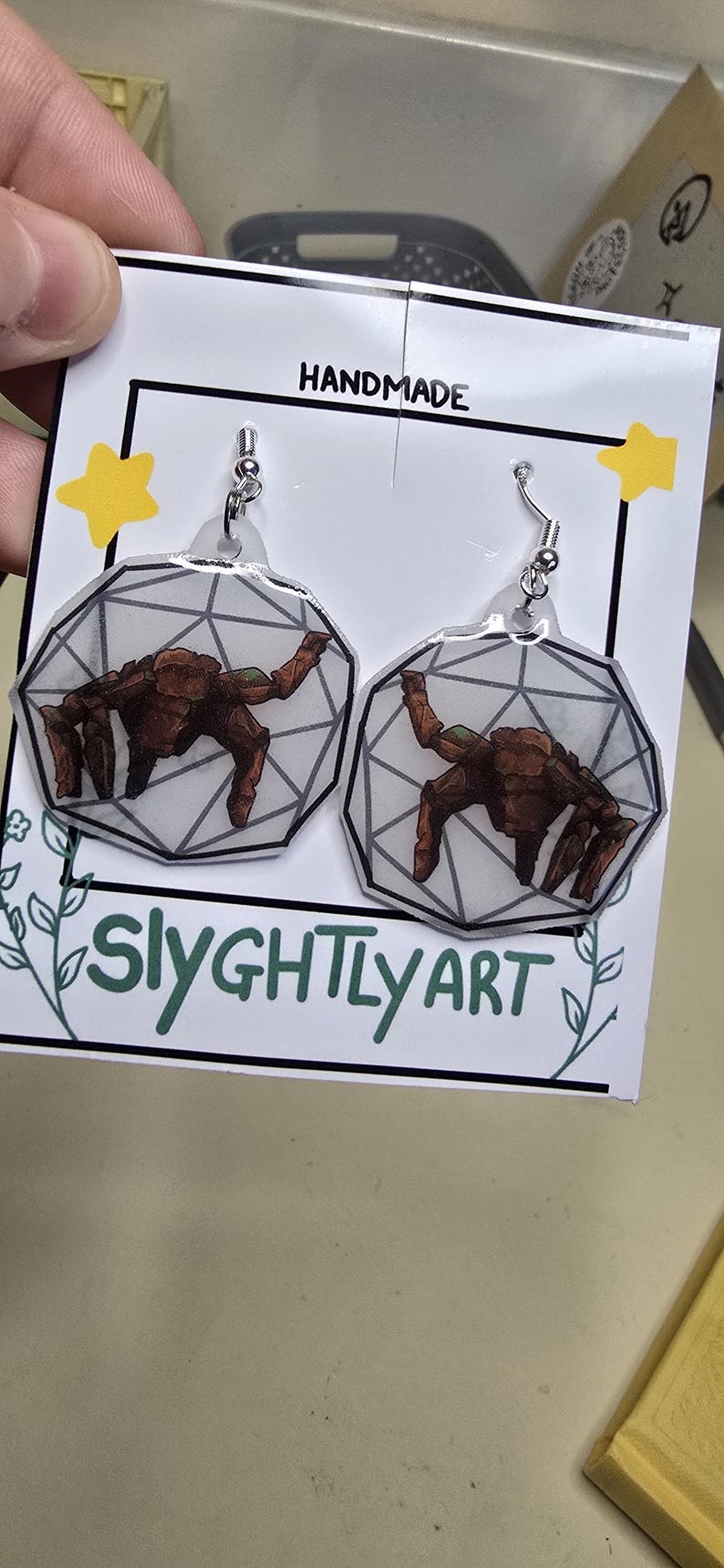May include: A pair of dangle earrings featuring a brown, multi-legged creature illustration inside a geometric, translucent, white, dodecahedron-shaped frame. The earrings are attached to a white display card labeled HANDMADE and SLYGHTLY ART.