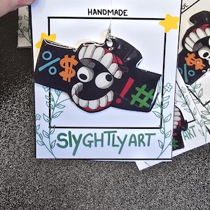 May include: Handmade black acrylic earrings with a cartoon face design. The earrings feature a smiling face with white teeth, red lips, and symbols like a percent sign, dollar sign, exclamation point, and hashtag. The card reads "HANDMADE" and "Slyghtly Art."