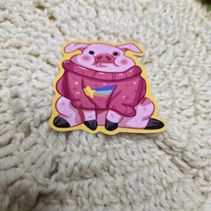 May include: A pink sticker featuring a cartoon pig wearing a pink sweater with a rainbow star. The pig is sitting down with its legs crossed. The sticker has a yellow border.