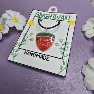 Peut inclure: Un collier fait à la main avec un pendentif en forme de tranche de pastèque, portant l'inscription "Viva la Vida". Le pendentif est suspendu à un cordon noir. Le produit est présenté sur une carte blanche avec "HANDMADE" et la marque "SlyghtlyArt".