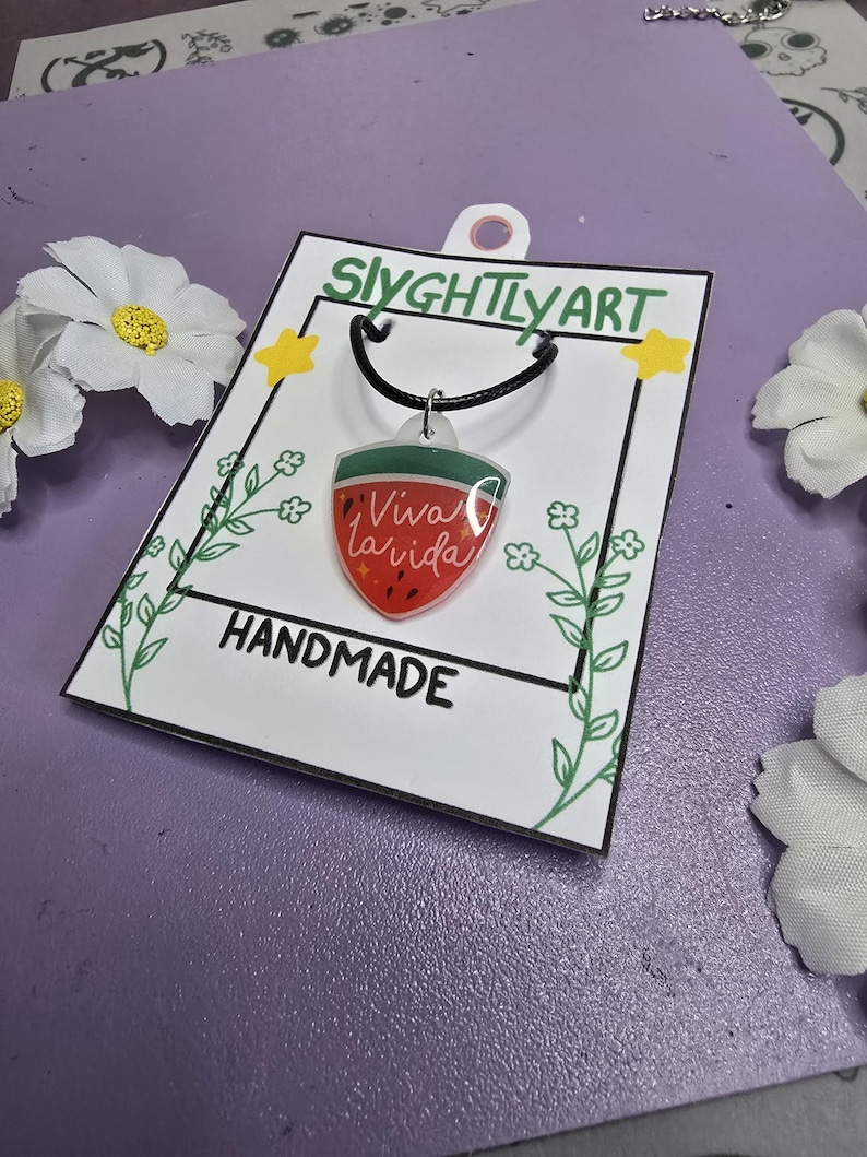 May include: A handmade necklace featuring a watermelon slice design with the words "Viva la Vida." The pendant hangs from a black cord. The product is displayed on a white card with the text "HANDMADE" and the brand name "SlyghtlyArt."