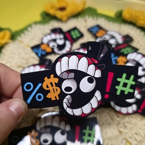 May include: Black sticker featuring a cartoon face with a top hat, large white teeth, and a wide grin. The sticker includes symbols like a percent sign, dollar sign, exclamation point, and a hashtag. The background is a crocheted surface.