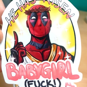 May include: A sticker featuring a cartoon depiction of Deadpool wearing a crown of thorns and a red and black suit. The sticker has a yellow background with the text "He Has Risen" in a pink circle around the character. Below the character is the text "Babygirl (F*ck!)" in pink and black.