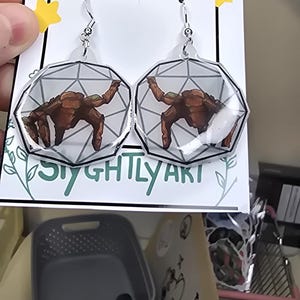 May include: A pair of dangle earrings featuring a brown, rocky creature illustration inside a clear, geometric, multi-faceted shape. The earrings are attached to a white display card with the text HANDMADE and SIGHTLY ART printed on it.