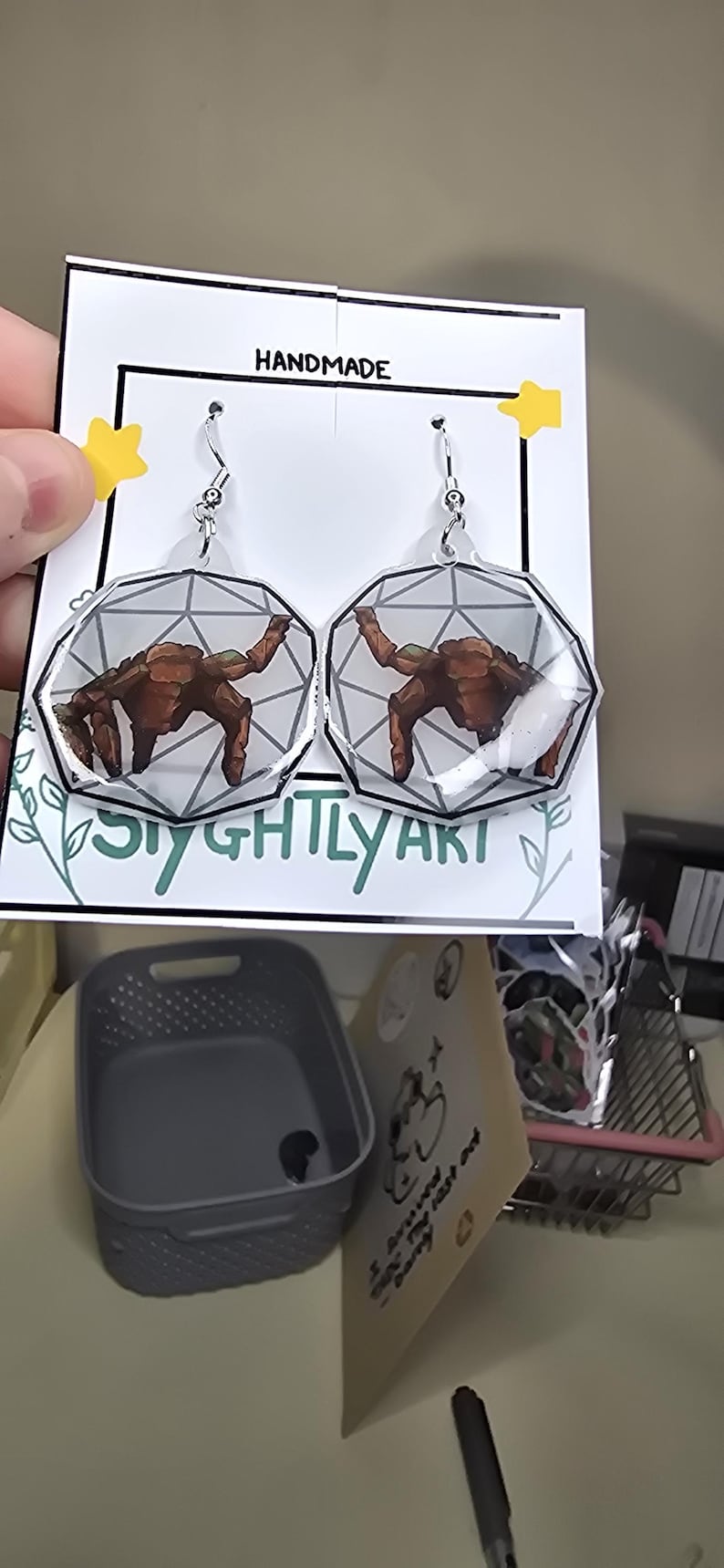 May include: A pair of dangle earrings featuring a brown, rocky creature illustration inside a clear, geometric, multi-faceted shape. The earrings are attached to a white display card with the text HANDMADE and SIGHTLY ART printed on it.