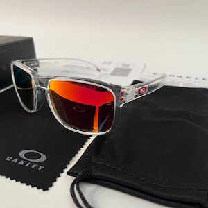 May include: Clear frame sunglasses featuring reflective orange lenses with a red tint. The temples display a small red Oakley logo. Included are a black microfiber cleaning cloth with the white Oakley logo and a black drawstring storage pouch.