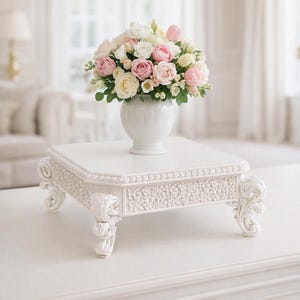 May include: A white square pedestal stand with ornate carved scroll legs and a textured floral border design. A white ceramic vase sits on top of the stand, filled with a lush arrangement of pink and white roses and small green foliage.