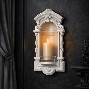 May include: A white wall-mounted candle niche featuring an arched top with decorative molding and a central shelf. A cylindrical cream-colored pillar candle sits inside a clear glass holder on the shelf, with a lit flame emitting a warm glow.