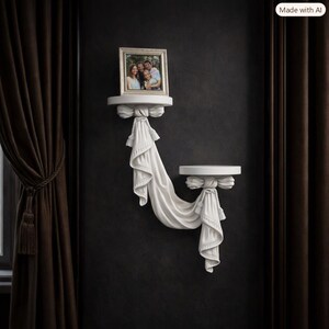 May include: A white wall-mounted shelf unit featuring two circular platforms connected by a sculpted fabric-like drape with tassels. The upper shelf holds a rectangular silver picture frame containing a photograph of three people.
