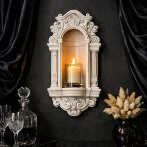May include: A cream-colored wall-mounted candle niche featuring an arched top, fluted columns, and ornate scrollwork detailing. A clear glass cylinder holds a single lit white pillar candle, providing a warm glow within the architectural frame.