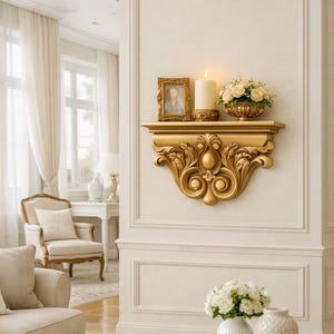 May include: A gold wall-mounted shelf featuring ornate carved scrollwork and acanthus leaf details. The shelf surface holds a small rectangular gold picture frame, a tall cylindrical cream-colored candle, and a gold bowl filled with white roses.