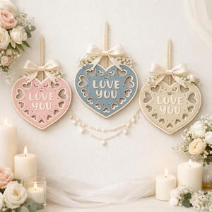 May include: Three heart-shaped wooden wall hangings in pastel pink, blue, and beige. Each heart features cut-out heart patterns, the text "LOVE YOU" in raised letters, a cream-colored satin ribbon bow, and small white floral accents.