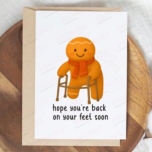 May include: A get-well card featuring a smiling gingerbread man with an orange scarf and a walker. The card has the text "hope you're back on your feet soon" in a black, handwritten font. The card is on a wooden tray.