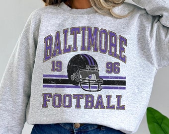 May include: A gray sweatshirt with a vintage-style graphic of a football helmet and the text "BALTIMORE 19 96 FOOTBALL" in purple and gold.