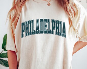 May include: A cream colored oversized t-shirt with the text "PHILADELPHIA" in dark green letters with a white outline. The shirt is being worn by a person with long blonde hair.
