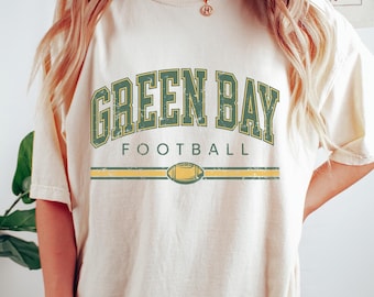 May include: A cream colored t-shirt with a vintage-style graphic that reads "Green Bay Football" in green and yellow lettering. The graphic includes a stylized football.