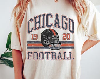 May include: Cream-colored t-shirt with a distressed graphic featuring the word "CHICAGO" above a football helmet and the word "FOOTBALL". The design includes the years "19" and "20" and is printed in navy blue, red, and white.