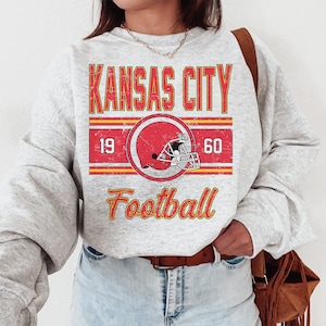 May include: A gray sweatshirt with a vintage-style graphic design featuring the text "KANSAS CITY FOOTBALL" with a red and white football helmet graphic. The text "19" and "60" are on either side of the helmet graphic.