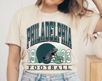 May include: A beige t-shirt with a vintage-style graphic design featuring the text "Philadelphia 19 33 Football" and a green and white football helmet.
