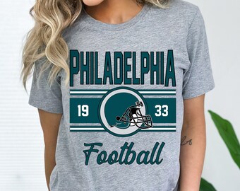 May include: Gray t-shirt with a teal green graphic of a football helmet and the text "PHILADELPHIA" with the numbers "19" and "33" on either side of the helmet. The text "Football" is below the graphic.