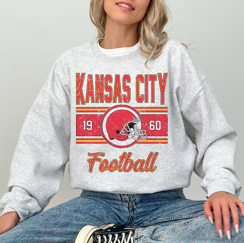 May include: A gray sweatshirt with a vintage-style graphic design featuring the text "Kansas City Football" with a red and white football helmet graphic and the numbers "19" and "60" on either side of the helmet.