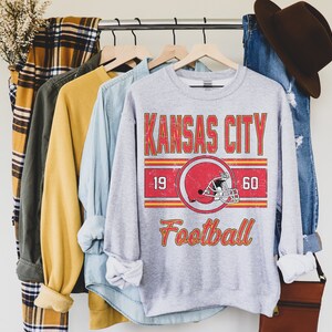May include: A gray sweatshirt with a vintage-style graphic design featuring the text "Kansas City Football" with a red and yellow color scheme. The graphic also includes a red football helmet with a white stripe.