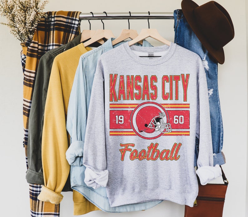 May include: A gray sweatshirt with a vintage-style graphic design featuring the text "Kansas City Football" with a red and yellow color scheme. The graphic also includes a red football helmet with a white stripe.