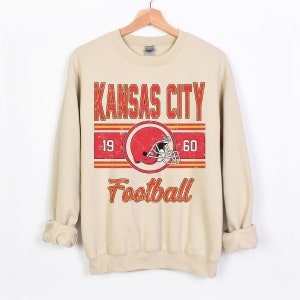 May include: A beige sweatshirt with a vintage-style graphic of the Kansas City Chiefs logo, the words "Kansas City Football", and the numbers "19" and "60".