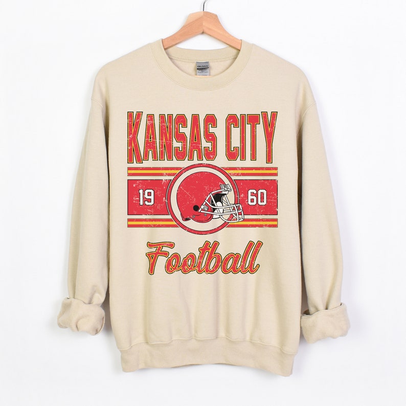 May include: A beige sweatshirt with a vintage-style graphic of the Kansas City Chiefs logo, the words "Kansas City Football", and the numbers "19" and "60".