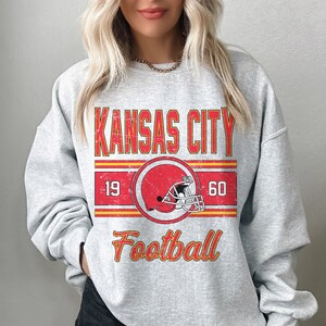 May include: Gray sweatshirt with a vintage-style graphic design featuring the text "KANSAS CITY" in red and yellow, a red football helmet, and the text "Football" in yellow. The numbers "19" and "60" are on either side of the helmet.