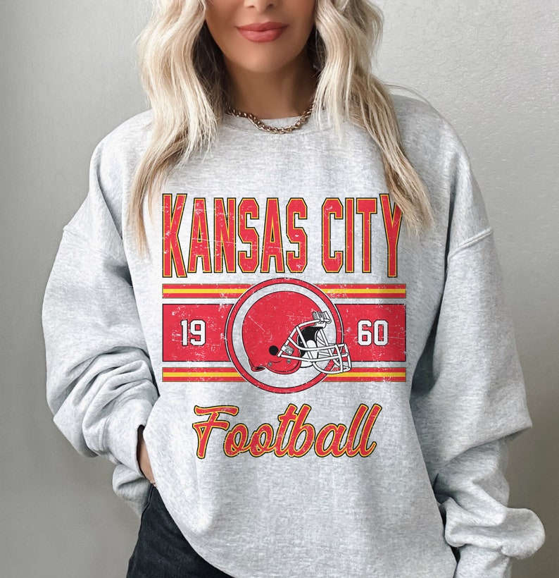 May include: Gray sweatshirt with a vintage-style graphic design featuring the text "KANSAS CITY" in red and yellow, a red football helmet, and the text "Football" in yellow. The numbers "19" and "60" are on either side of the helmet.