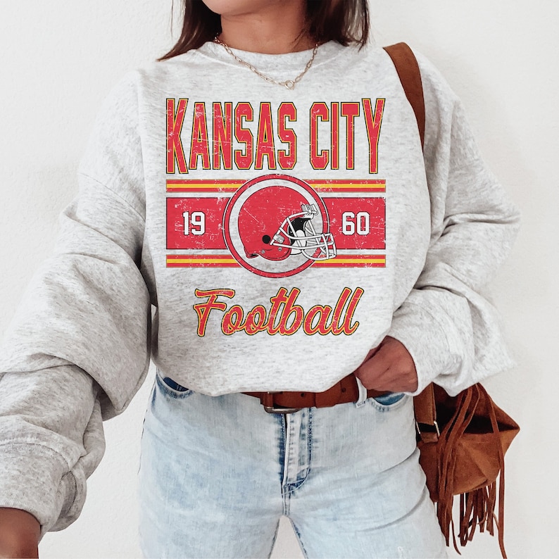 May include: A gray sweatshirt with a vintage-style graphic design featuring the text "KANSAS CITY FOOTBALL" with a red and white football helmet graphic. The text "19" and "60" are on either side of the helmet graphic.