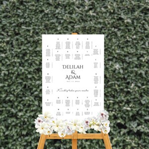 Op de afbeelding: Een wit huwelijkszitplan met zwarte tekst. Het plan staat op een ezel en heeft de namen "Delilah & Adam" en de datum "May 11, 20xx". Het plan is versierd met witte bloemen.