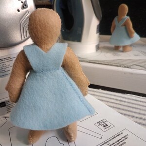 May include: A light blue felt doll with a brown felt head and arms. The doll is wearing a light blue dress with a V-neckline. The doll is standing on a white surface.